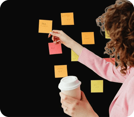 Personne en veste rose collant des notes adhésives colorées sur un mur noir, tenant un gobelet de café